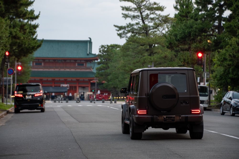 京都・平安神宮の朱塗りの社殿を背に、神宮道を走るマットブラウンのメルセデス・ベンツ Gクラスのリアビュー（FOURSIDE）